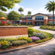 Realistic photograph of a well-maintained school campus with a two-story brick building, curved walkway leading to the entrance, landscaped gardens with colorful flowers, tall palm trees, and a playground structure in the background under a clear blue sky.